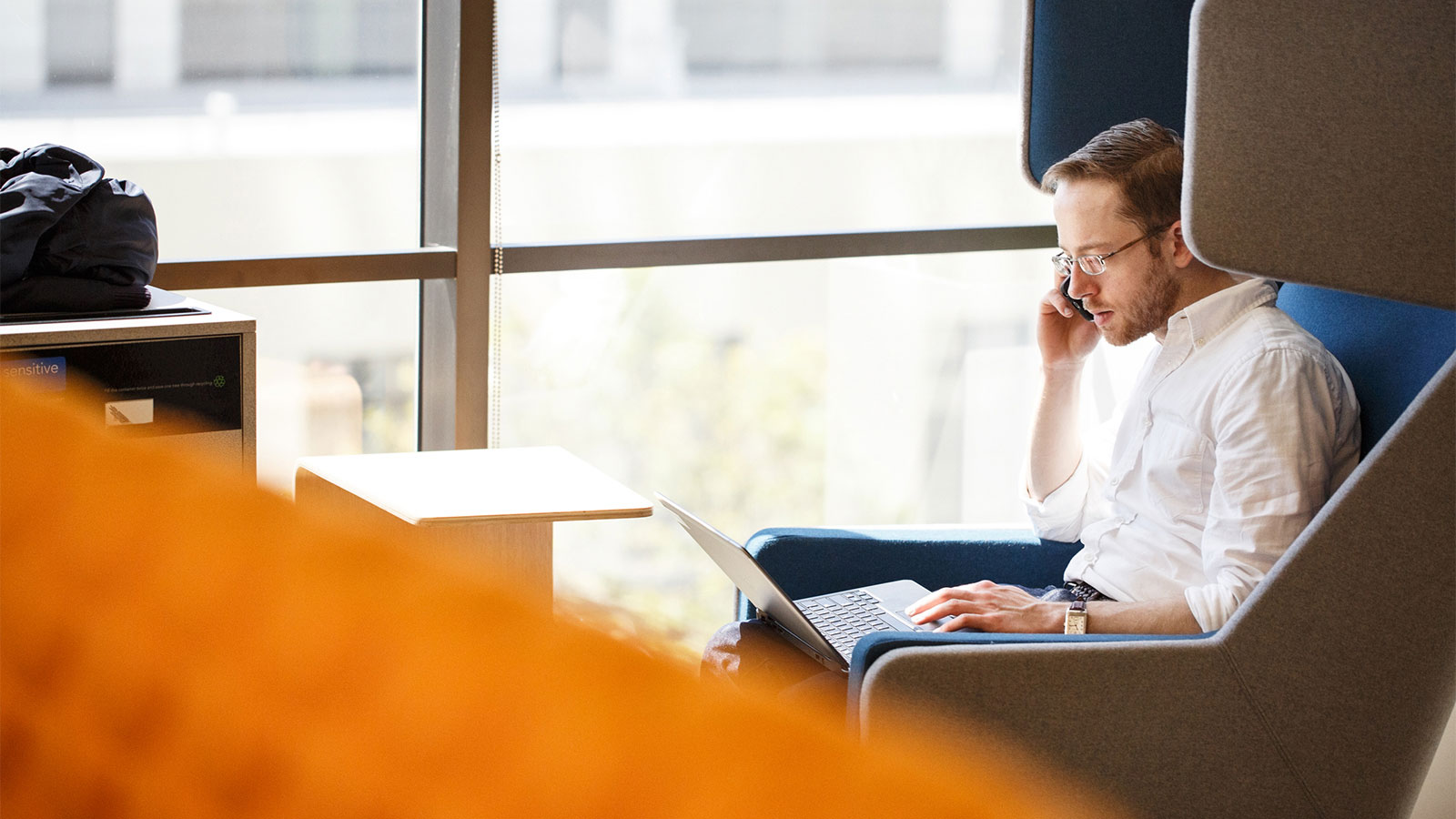 Man sitting on a chair in office environment, talking on his cell phone while looking at his laptop.